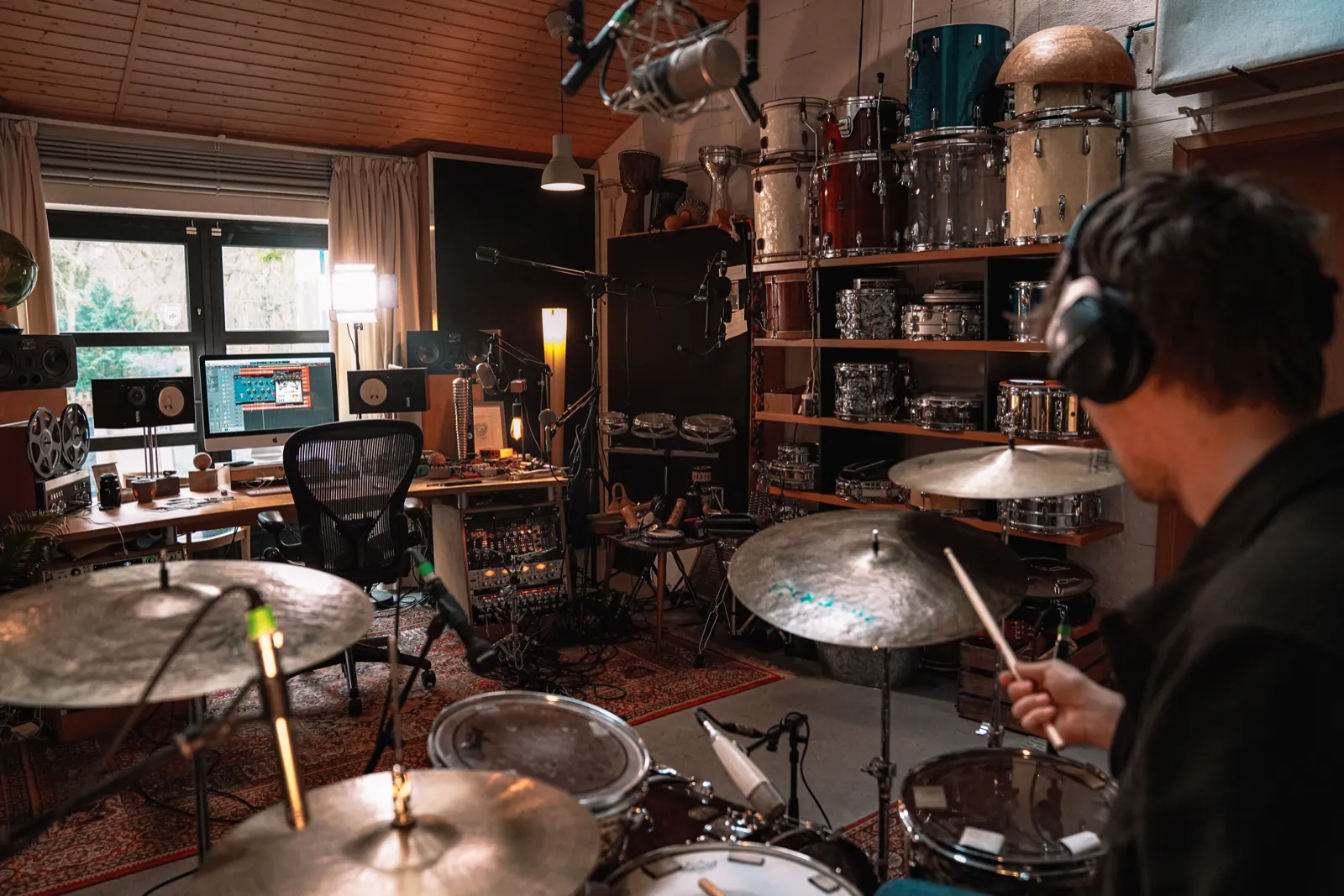 A drummer with headphones playing a miked-up kit in his recording room, shelves of snare drums and a computer desk behind him