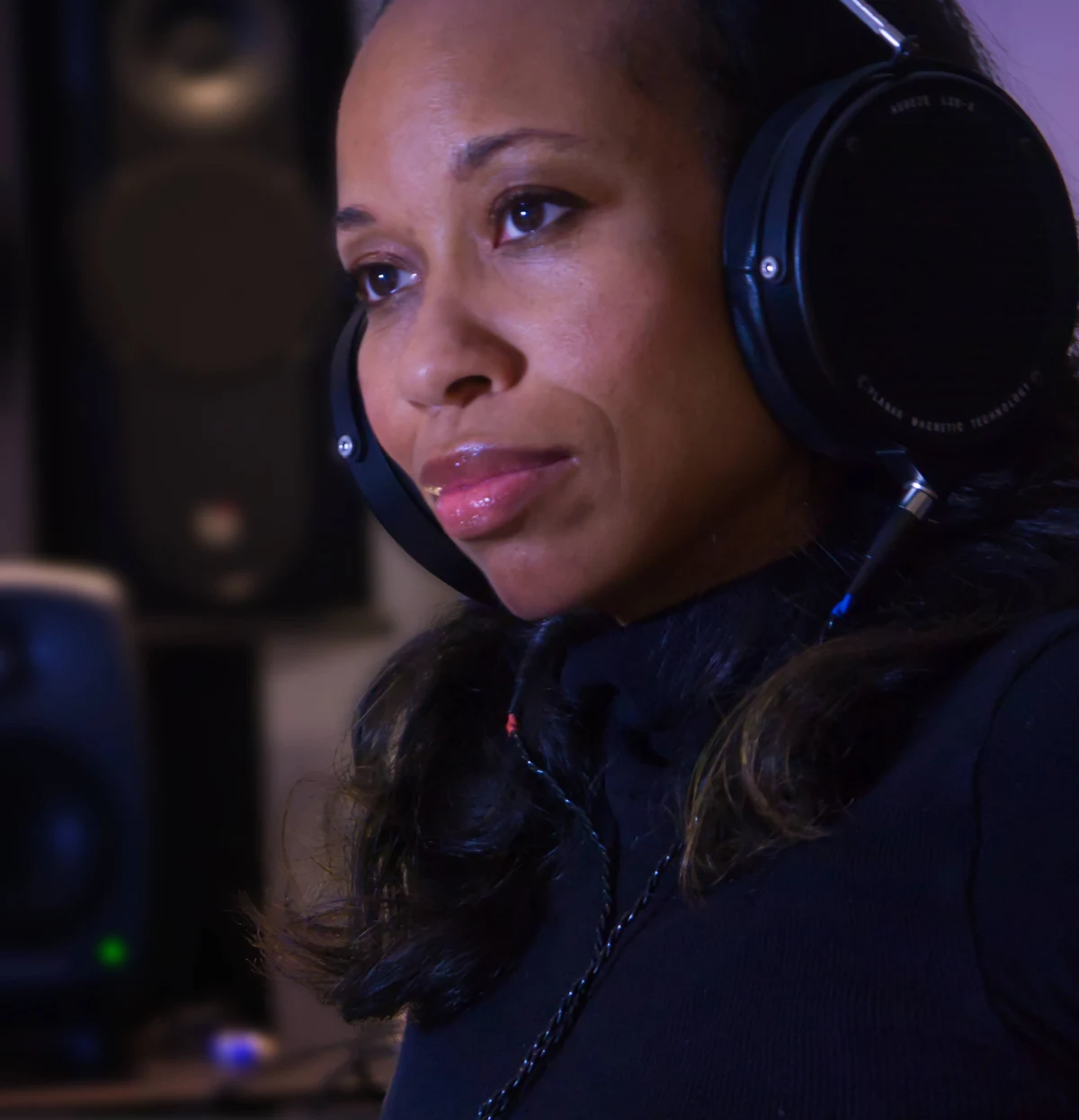 A woman wearing open-back headphones, listening to an Atmos mix in her studio, two monitor speakers behind her